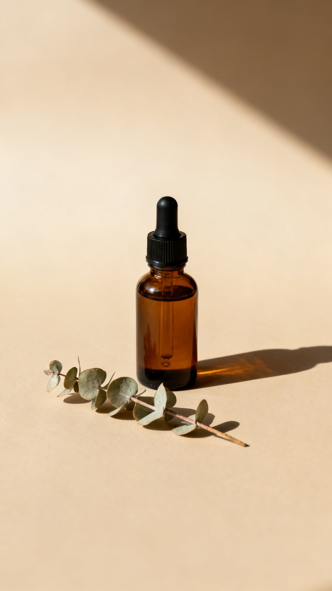 An amber glass dropper bottle on a beige background beside a eucalyptus sprig
